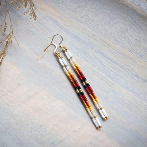 Puede incluir: Un par de pendientes largos de cuentas con un degradado de cuentas rojas, naranjas, amarillas, blancas y negras. Los pendientes están hechos con cuentas pequeñas y redondas y tienen ganchos de color dorado.