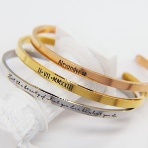 Puede incluir: Tres pulseras de brazalete apiladas en oro, plata y oro rosa con texto grabado. La pulsera superior dice "Alexander" con una huella de pata. La pulsera del medio dice "II. VII. MMXXIII". La pulsera inferior dice "Deja que la belleza de lo que amas sea lo que haces."