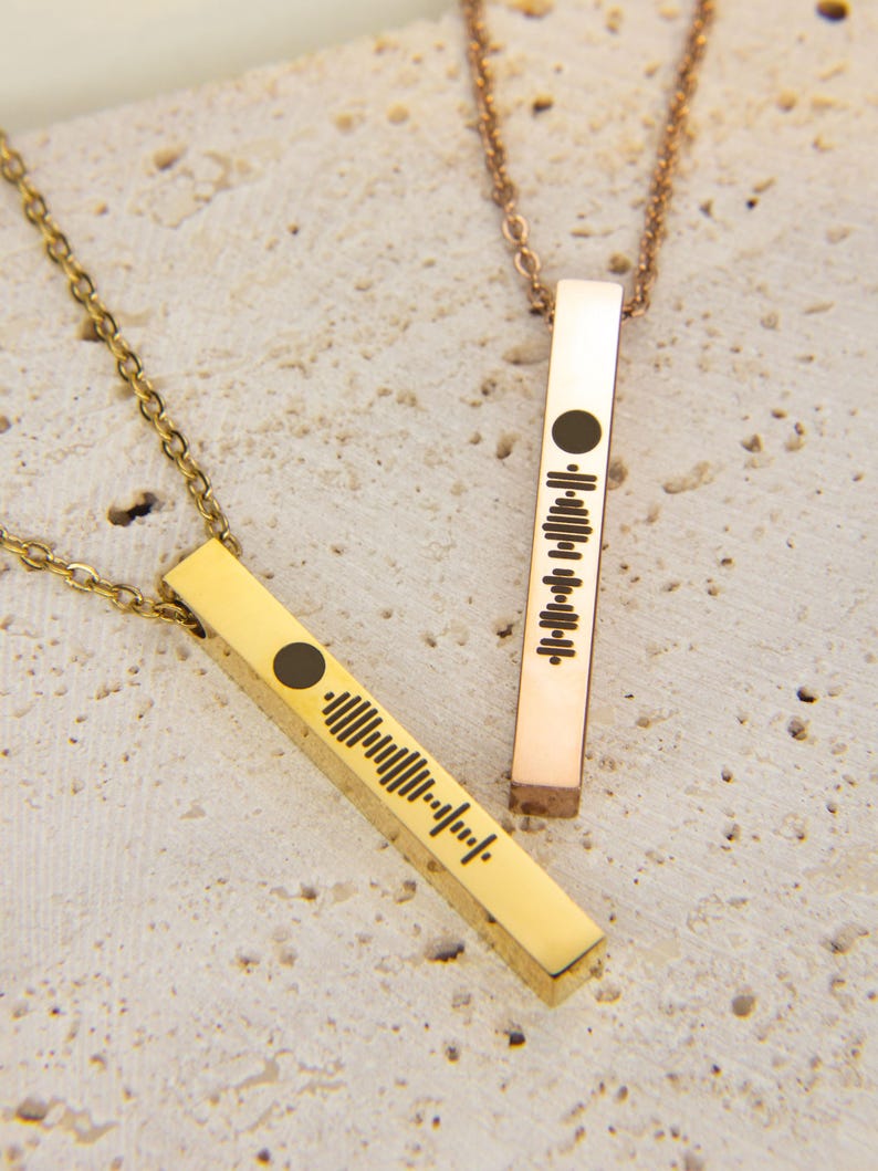 May include: Two gold bar necklaces with a soundwave design. The necklace on the left is gold and the necklace on the right is rose gold.