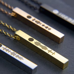 May include: Four gold bar necklaces with a black dot and a barcode-like design. The necklaces are on a dark grey surface.