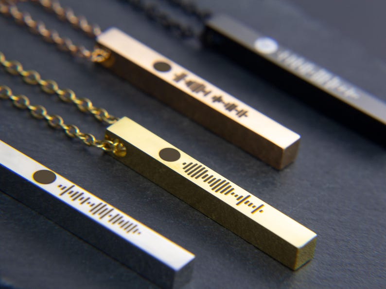 May include: Four gold bar necklaces with a black dot and a barcode-like design. The necklaces are on a dark grey surface.