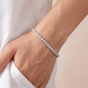 以下が含まれることがあります： 金属製のフレームにセットされた、丸く透明なファセットカットの石が連なるシルバー調のテニスブレスレット。ブレスレットは手首に装着されており、輝く円形の宝石が一直線に並んだ均一なデザインが特徴です。