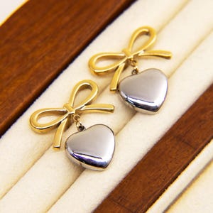 Puede incluir: Un par de pendientes de lazo dorado con un colgante de corazón plateado que cuelga de cada lazo.