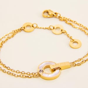 May include: A gold-tone bracelet with a double chain design. The bracelet features a large, round pendant with a white mother-of-pearl inlay and a small clear crystal. The pendant has the word "Forever" engraved on it.