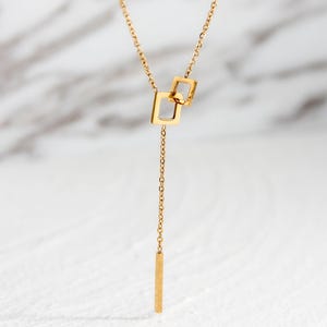 Puede incluir: Un collar de cadena dorada con un colgante que presenta dos formas cuadradas entrelazadas y una barra larga y delgada que cuelga de la parte inferior.