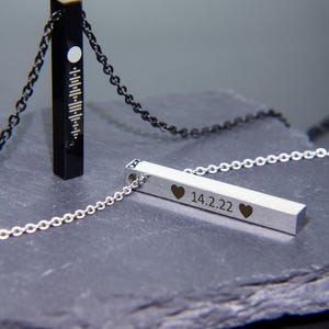 May include: Two silver bar necklaces with chains. One necklace has a black bar with a white circle and lines. The other necklace has a silver bar with the inscription "14.2.22" and two hearts.