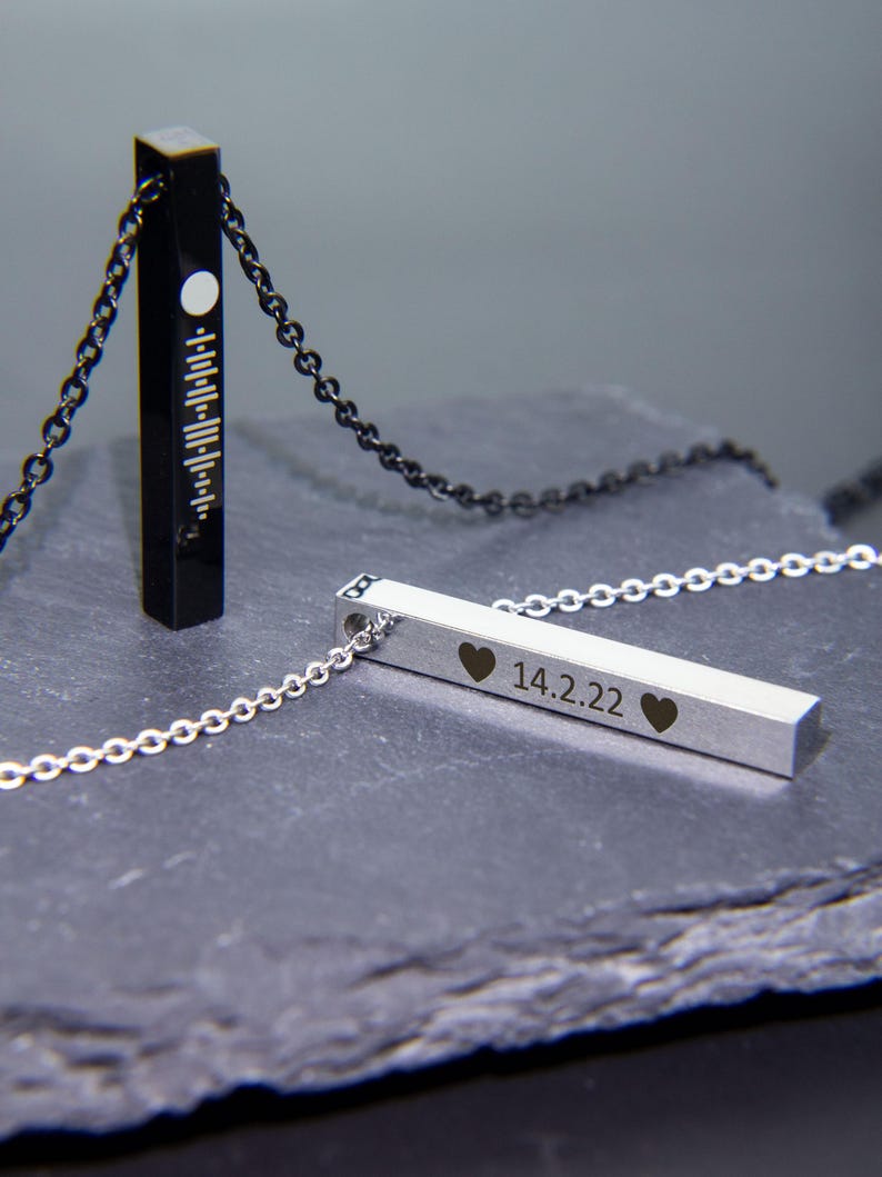 May include: Two silver bar necklaces with chains. One necklace has a black bar with a white circle and lines. The other necklace has a silver bar with the inscription "14.2.22" and two hearts.