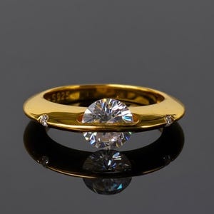 Puede incluir: Un anillo dorado con una gema grande, clara y redonda en el centro. El anillo tiene una banda ancha y pequeñas piedras de acento. El anillo se refleja en la superficie oscura de abajo. El anillo está marcado con "S925".