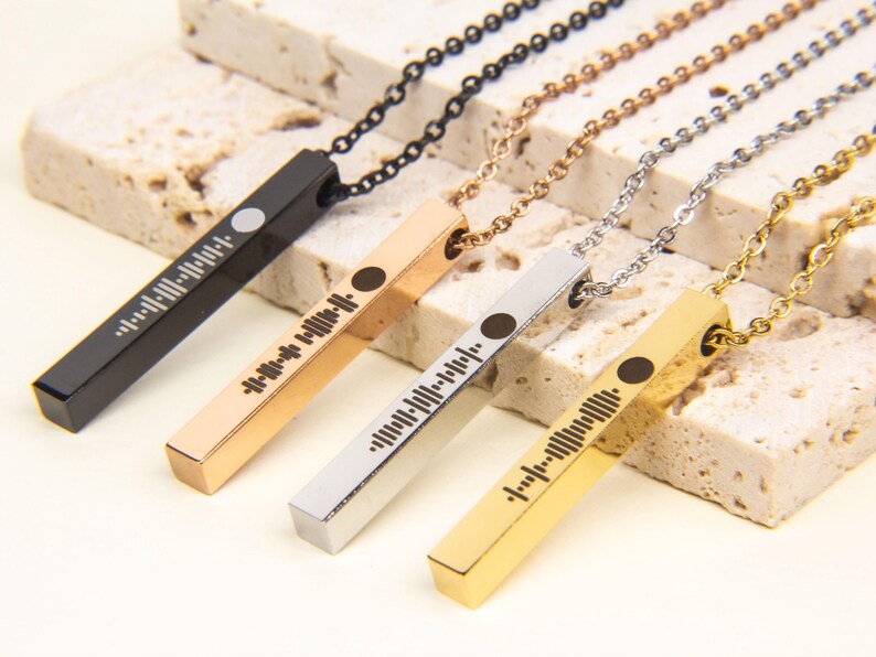 May include: Four bar necklaces with different coloured metal finishes: black, rose gold, silver, and gold. Each bar has a unique barcode-like design engraved on it.