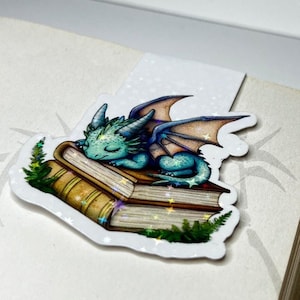 Könnte beinhalten: Ein blaues und grünes Drachen-Lesezeichen mit Glitzer, das auf einem Stapel Bücher schläft. Das Lesezeichen ist weiß mit einem weißen Rand und hat einen weißen Hintergrund. Der Text "SKY ABOVE, EARTH BELOW, PEACE WITHIN" ist auf der Unterseite des Lesezeichens gedruckt.