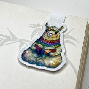 Könnte beinhalten: Ein skurriles Lesezeichen mit einer farbenfrohen Lama-Illustration. Das Lama trägt einen Regenbogen-Schal und hält ein Buch. Das Lesezeichen ist an einem Buch befestigt. Das Fineline Mountains Logo ist sichtbar.