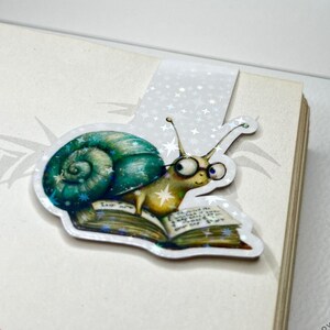 Op de afbeelding: Een glinsterende witte bladwijzer met een slak die een bril draagt en op een boek zit. De slak is groen en goud met een blauwe schelp. De bladwijzer heeft een witte achtergrond met zilveren glittersterren.