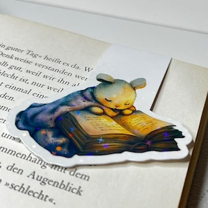 Könnte beinhalten: Ein weißes Kaninchen, das eine blaue Decke trägt, liest ein Buch mit goldenem Einband und funkelnden Sternen. Das Buch ist geöffnet und das Kaninchen liegt darauf zusammengerollt. Das Kaninchen hat einen süßen Gesichtsausdruck und genießt das Buch sichtlich.