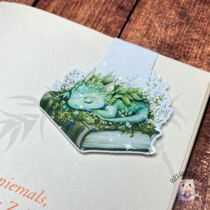 Op de afbeelding: Een fantasievolle boekenlegger met een slapende groene draak op een boek. De draak is versierd met bladeren en rust te midden van bloemendetails. De boekenlegger heeft een glinsterende, iriserende afwerking.