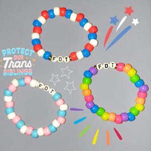 May include: Three beaded bracelets with the letters "F.D.T." spelled out on each. The bracelets are in the colors of the American flag, the transgender pride flag, and the rainbow pride flag.