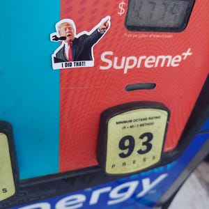 May include: A sticker of a man pointing, with the text "I DID THAT!" is placed on a red and blue gas pump. The pump displays a price of $4.779 per gallon. The brand "Supreme+" is visible.
