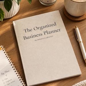 May include: A beige business planner with the title The Organized Business Planner by Rebecca LaBranch printed in black text on the cover. The planner is placed next to a spiral-bound notebook with a Big Plans checklist and a white pen.