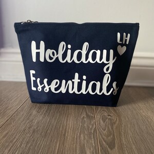 May include: A navy blue canvas cosmetic bag with a silver zipper. The words "Holiday Essentials" are printed in white script across the front. The initials "LH" and a heart are printed on the upper right corner.