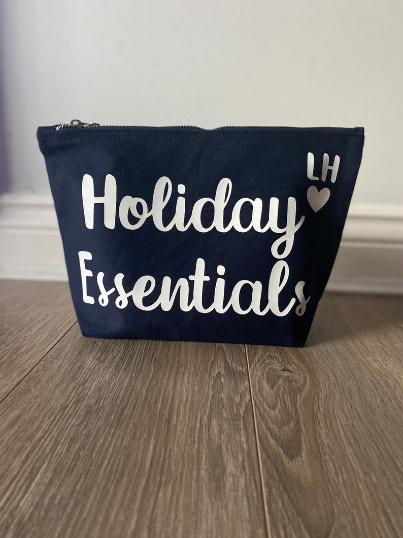 May include: A navy blue canvas cosmetic bag with a silver zipper. The words "Holiday Essentials" are printed in white script across the front. The initials "LH" and a heart are printed on the upper right corner.