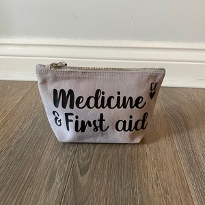 May include: A light gray canvas pouch with a silver zipper. The words "Medicine & First aid" are printed in black script on the front. Two small black hearts are printed next to the words. The pouch is on a wooden surface.
