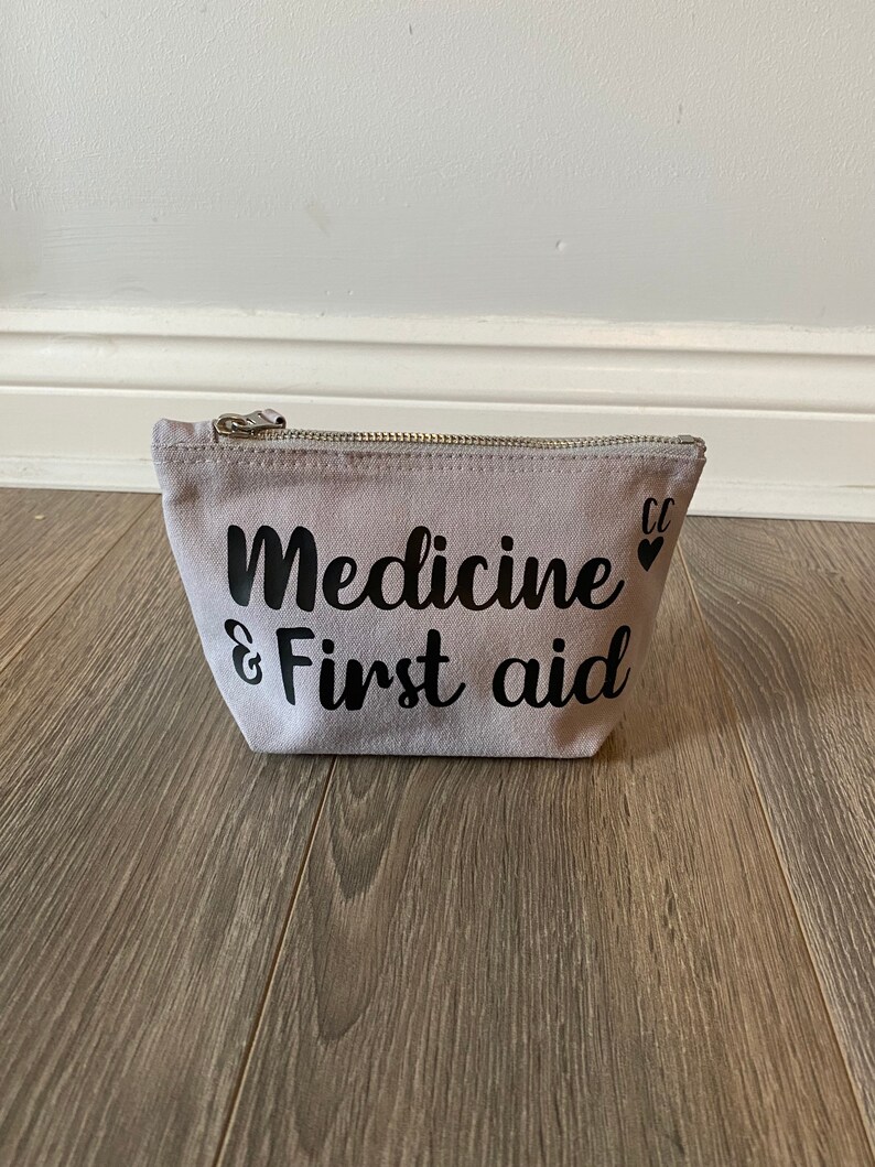 May include: A light gray canvas pouch with a silver zipper. The words "Medicine & First aid" are printed in black script on the front. Two small black hearts are printed next to the words. The pouch is on a wooden surface.