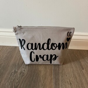 May include: A light gray canvas zippered pouch with the words "Random Crap" in black script. The initials "CC" and a heart are printed on the upper right corner. The pouch is sitting on a wooden floor.