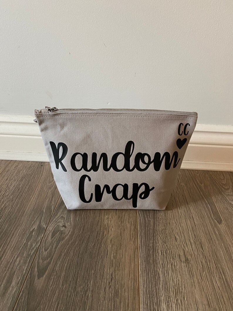 May include: A light gray canvas zippered pouch with the words "Random Crap" in black script. The initials "CC" and a heart are printed on the upper right corner. The pouch is sitting on a wooden floor.