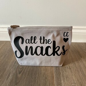 May include: A light gray canvas zippered pouch with the phrase "all the snacks" in black script. The pouch has a small heart and the initials "CC" printed on the right side.