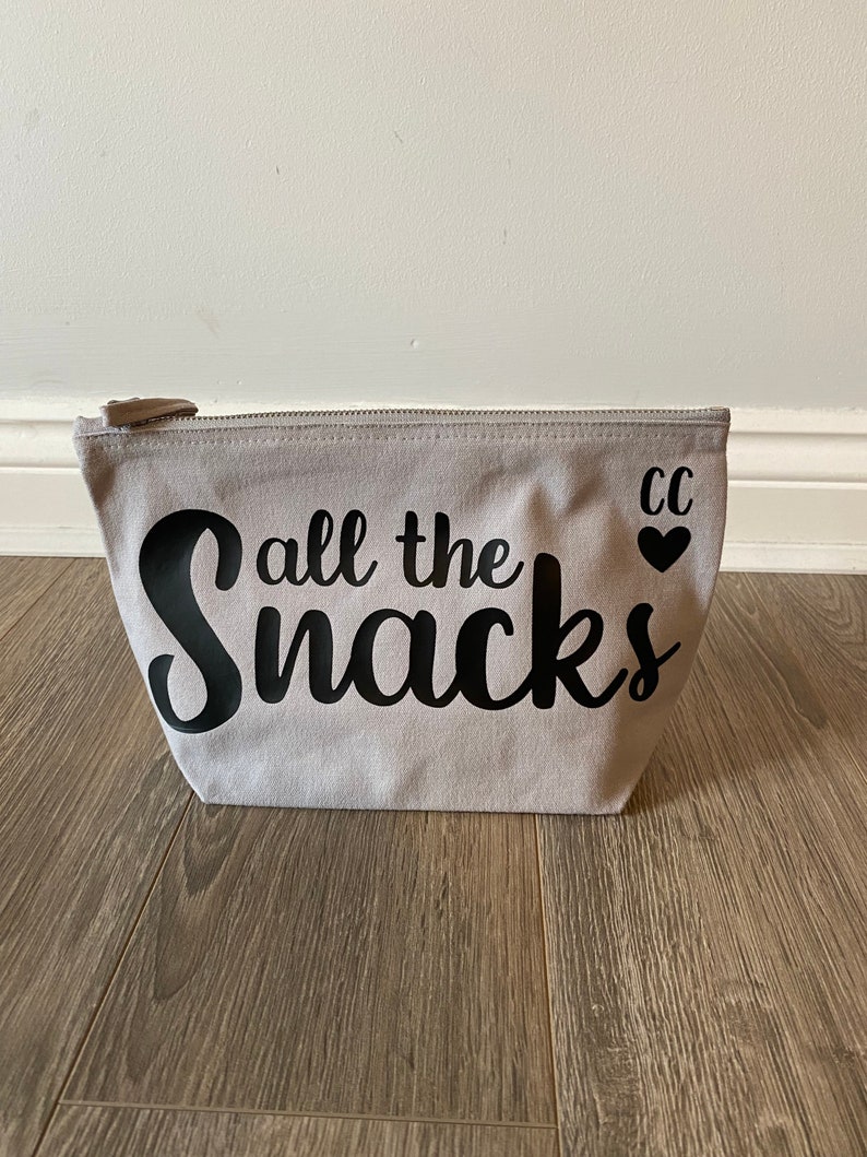 May include: A light gray canvas zippered pouch with the phrase "all the snacks" in black script. The pouch has a small heart and the initials "CC" printed on the right side.