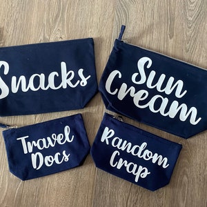 May include: Four navy blue zippered pouches with white text. The pouches are labeled "Snacks", "Sun Cream", "Travel Docs", and "Random Crap". The pouches are rectangular and made of a canvas-like material.