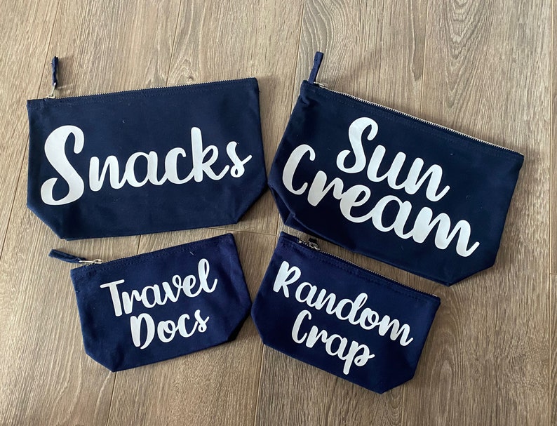 May include: Four navy blue zippered pouches with white text. The pouches are labeled "Snacks", "Sun Cream", "Travel Docs", and "Random Crap". The pouches are rectangular and made of a canvas-like material.