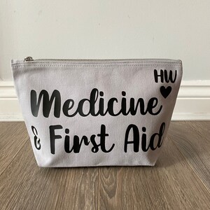 May include: A light gray canvas cosmetic bag with a silver zipper. The words "Medicine & First Aid" are printed in black script. The initials "HW" and a heart are printed on the right side of the bag.
