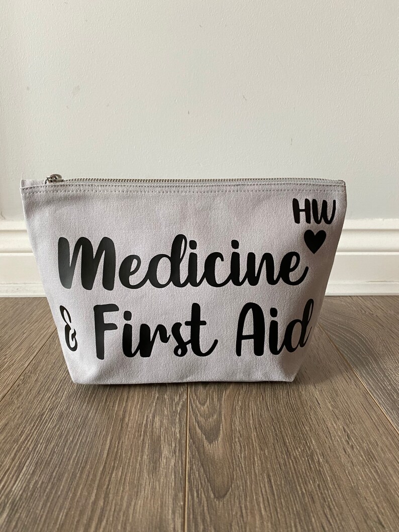 May include: A light gray canvas cosmetic bag with a silver zipper. The words "Medicine & First Aid" are printed in black script. The initials "HW" and a heart are printed on the right side of the bag.