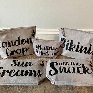 May include: A set of five gray canvas zipper pouches with black text. The pouches read "Random Crap", "Medicine & First aid", "Bikinis", "Sun Creams", and "all the Snacks". Each pouch has a silver zipper and a small black heart.