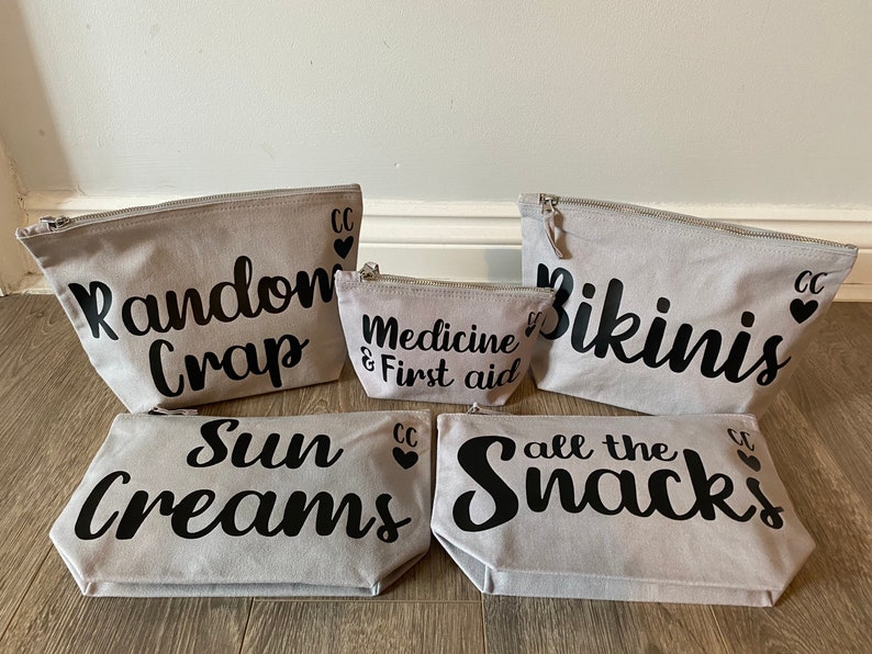 May include: A set of five gray canvas zipper pouches with black text. The pouches read "Random Crap", "Medicine & First aid", "Bikinis", "Sun Creams", and "all the Snacks". Each pouch has a silver zipper and a small black heart.