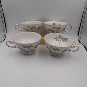 May include: Four white teacups with a gold rim and a green leaf design. The cups are arranged in a square formation.