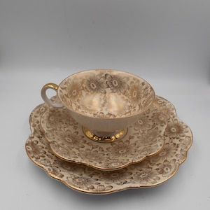 May include: A vintage teacup and saucer set with a delicate floral pattern in gold on a cream background. The set has a scalloped edge and is perfect for afternoon tea.