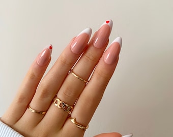 May include: Close-up of a hand with almond-shaped nails painted with a classic French manicure. The tips of the nails are painted white with a small red heart accent on each nail.