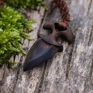 Puede incluir: Un collar con colgante de madera marrón oscuro con un diseño en forma de daga. El colgante está suspendido de un cordón trenzado marrón. El fondo presenta musgo verde y una superficie de madera envejecida.