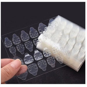 May include: A sheet of clear adhesive tabs for attaching false nails. The tabs are arranged in rows and columns on a white backing sheet. The sheet is partially rolled up, revealing more tabs underneath.