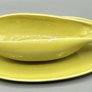 May include: A pale yellow ceramic gravy boat with a matching saucer. The gravy boat has a long, oval shape with a handle on one side. The saucer is round and flat. The set is made of ceramic.