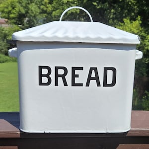 May include: A white metal bread box with a rectangular shape, featuring the word BREAD printed in bold black capital letters on the front. The container has a removable lid with a curved handle on top and small handles on both sides.