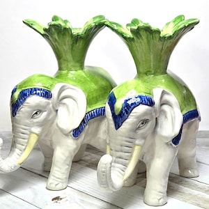 May include: Pair of ceramic elephant figurines. The elephants are white with green and blue accents. The elephant's backs are green, and the ears are white. The trunks are a light yellow. The top of the elephants have a green, leaf-like design.