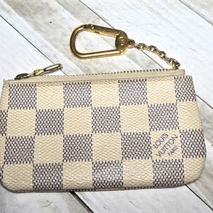 May include: A small, beige and brown checkered coin purse with a gold-colored zipper and key ring. The words "LOUIS VUITTON PARIS" are printed on the front of the purse. The key ring is attached to the purse with a small chain.