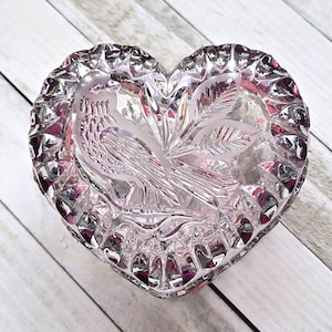 May include: Heart-shaped, clear glass trinket box with a bird and leaf design. The box has a faceted edge and a pinkish hue. The lid features an etched bird and leaf motif, adding a decorative touch to this small container.