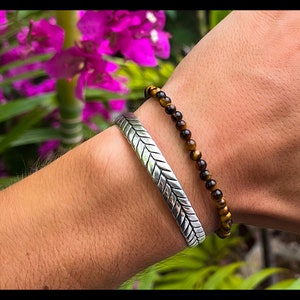Peut inclure: Un bracelet de manchette argenté avec un motif en chevron porté sur un poignet avec un bracelet de perles brun et or.