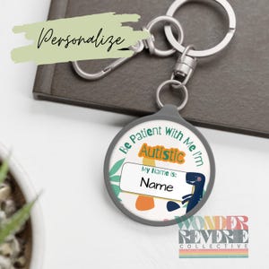 May include: A personalized keychain with a gray border and a round design. The design includes the text "Be Patient With Me I'm Autistic" and a space to write a name. The keychain also features a dinosaur illustration and the word "Personalize".