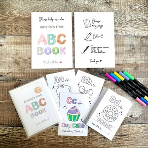 May include: A set of children's ABC coloring books with the text "Amelia's first ABC BOOK". The image includes coloring pages with illustrations of an apple, bow, cupcake, and donut. A set of colorful markers is also visible.