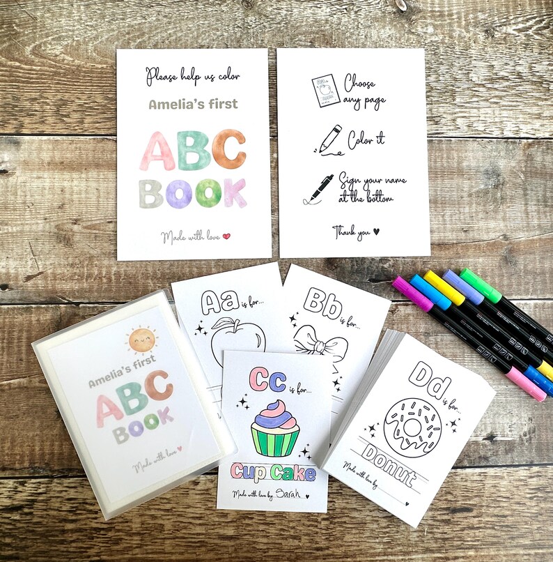 May include: A set of children's ABC coloring books with the text "Amelia's first ABC BOOK". The image includes coloring pages with illustrations of an apple, bow, cupcake, and donut. A set of colorful markers is also visible.