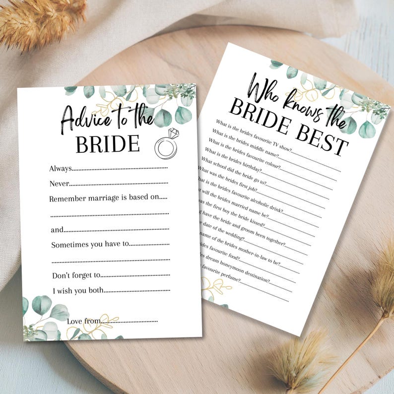 May include: Two white bridal shower game cards with eucalyptus and gold accents. One card reads "Advice to the Bride" and the other reads "Who Knows the Bride Best". The cards are on a wooden surface.